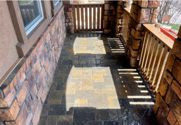 A sunlit stone-tiled porch with a wooden railing and gate, bordered by brick walls and a window on the left side.