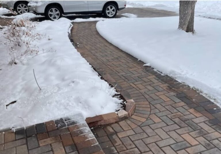 A brick walkway curves through a snow-covered yard toward a driveway where two cars are parked; snow is cleared from the walkway.