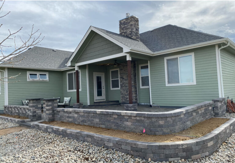 A single-story house with light green siding, a stone chimney, and a partially built stone patio surrounded by gravel and unfinished landscaping.