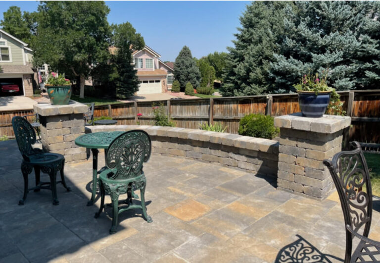Patio with green metal table and chairs, stone wall, potted plants, and a view of a suburban neighborhood with trees and houses in the background.
