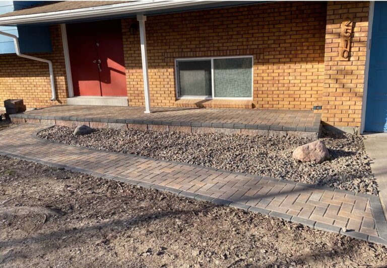 A brick-fronted house with a paved walkway and porch, bordered by rocks and gravel, with a bare dirt area in the foreground.