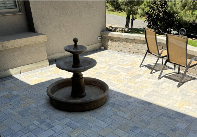 A stone patio with a tiered water fountain in the center and two tan patio chairs near a low brick wall, with trees and a parked car in the background.