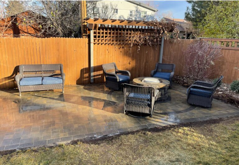 Outdoor patio with four wicker chairs, a loveseat, and a round fire pit on a paved surface. Wooden fence and trellis with sparse plants in the background.