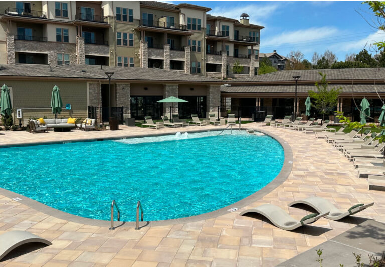 Outdoor swimming pool surrounded by lounge chairs and umbrellas, with apartment buildings and a clubhouse in the background on a sunny day.