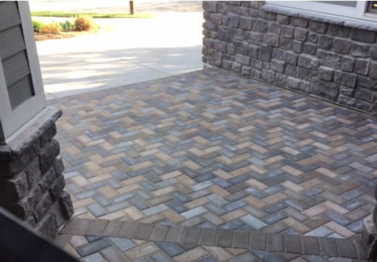 A patio with herringbone-patterned brick pavers bordered by stone walls and partial view of a driveway and landscaping in the background.