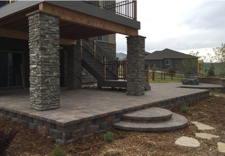 A covered stone patio with large stone columns, an upper deck with metal railings, and a set of curved steps leading to a landscaped yard.