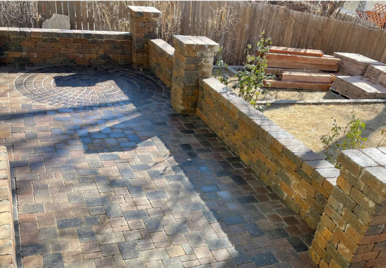 Stone patio with low retaining walls, a circular paver design, and wooden raised garden beds in a fenced backyard.