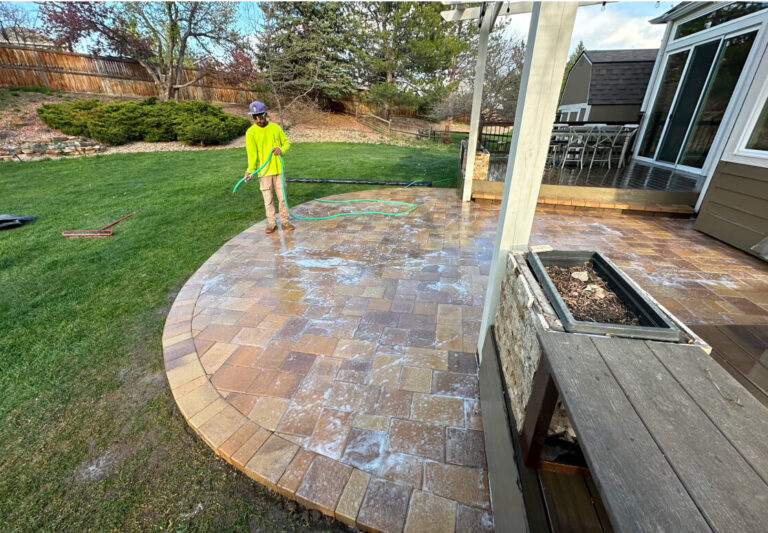 A person in a yellow shirt sprays water on a soapy stone patio with a garden hose near a backyard lawn and outdoor furniture.