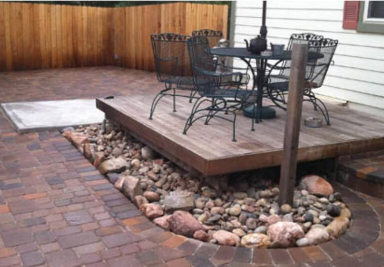 A small wooden deck with a metal table and chairs sits above a bed of rocks, surrounded by brick pavers in a backyard enclosed by a wooden fence.