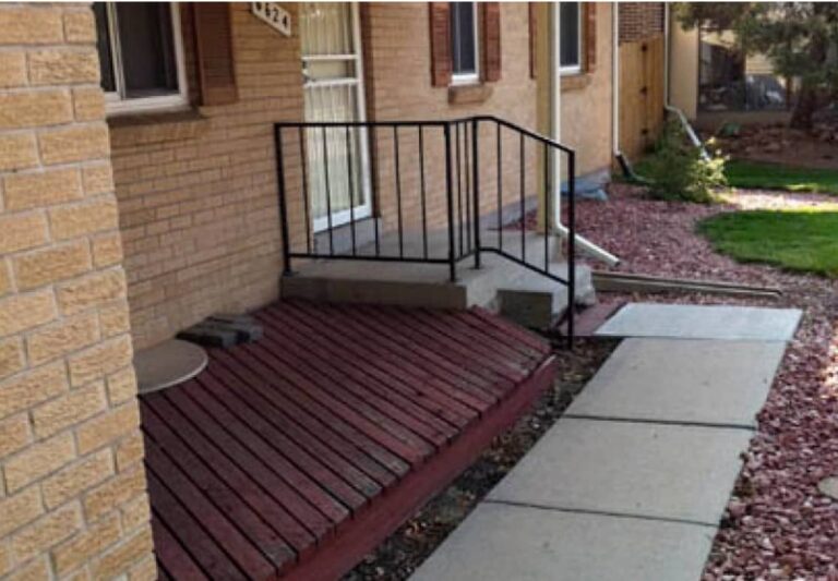 A sidewalk leads to a small front porch with a metal railing and steps. The porch is above a slatted wooden structure next to the house, surrounded by red gravel landscaping.