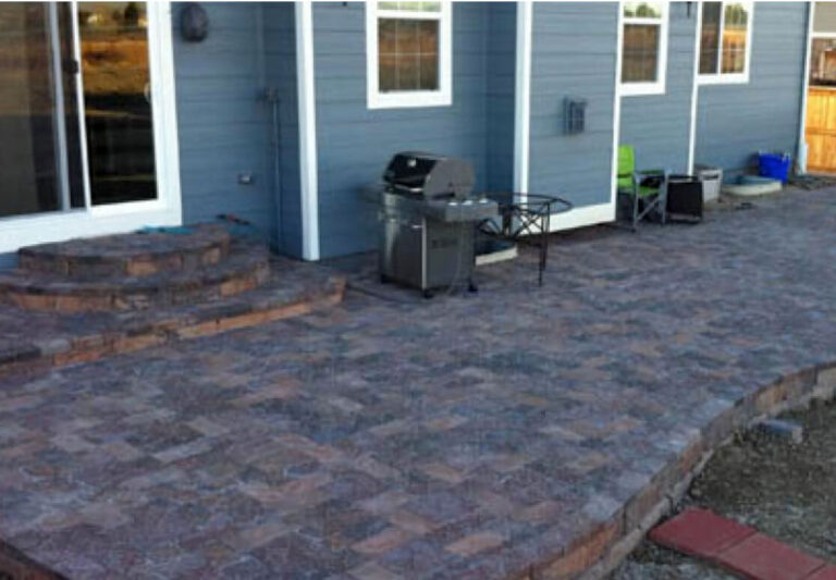 A backyard patio with stone pavers, a grill, a table, two chairs, and steps leading to a sliding glass door of a blue house.