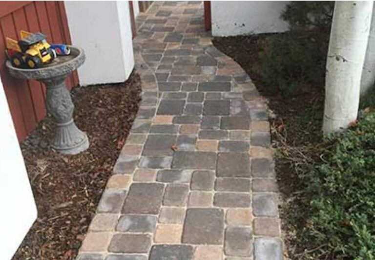 Curved stone paver walkway bordered by mulch and bushes, with a stone pedestal holding toy trucks on the left side.