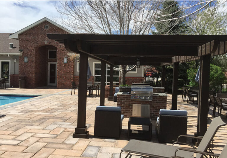 Outdoor patio with a brick house, pergola, grill, lounge chairs, dining table, and pool on a paved stone deck, surrounded by trees and greenery under a partly cloudy sky.