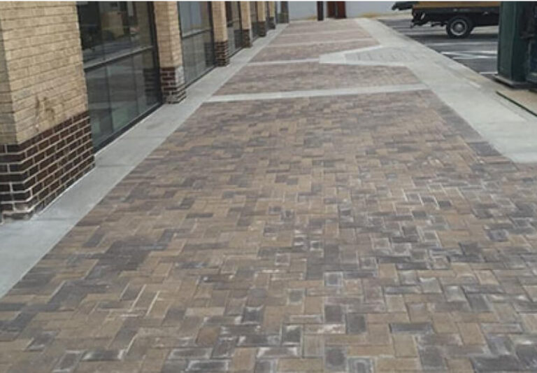 A newly paved sidewalk with rectangular bricks in a herringbone pattern runs alongside a building with large windows and brick walls.