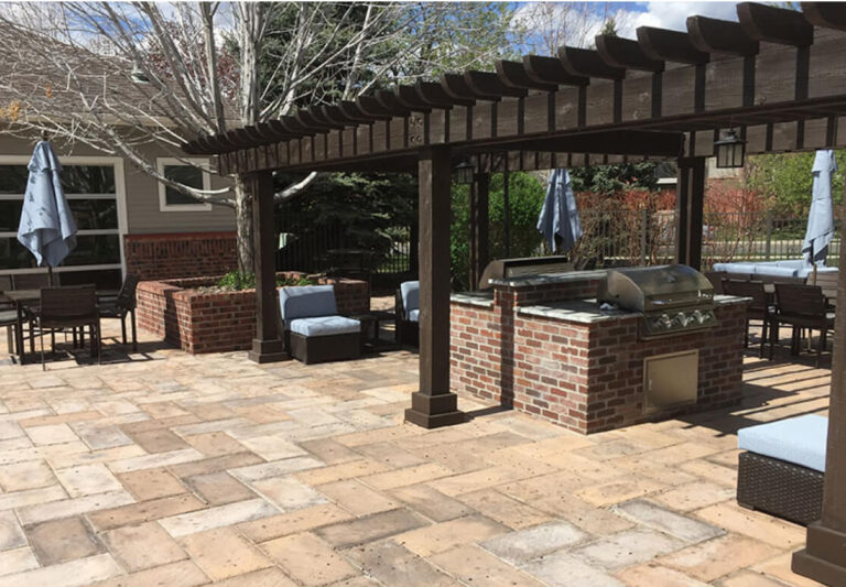 Outdoor patio with brick grill station under a wooden pergola, cushioned seating, tables with umbrellas, and stone tile flooring.