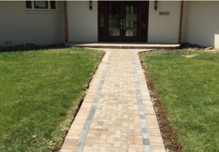 A paved stone walkway with gray accent bricks leads to a set of double front doors on a house, bordered by green grass.