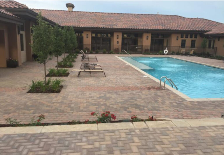 Rectangular outdoor swimming pool with poolside lounge chairs, surrounding paver deck, small landscaped areas, and a tile-roofed building in the background.