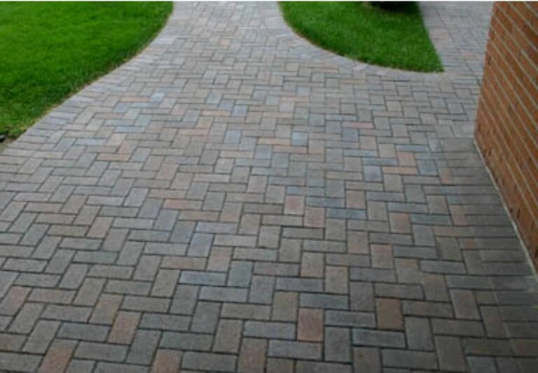 Curved walkway made of interlocking bricks, bordered by green grass and next to a brick building wall.
