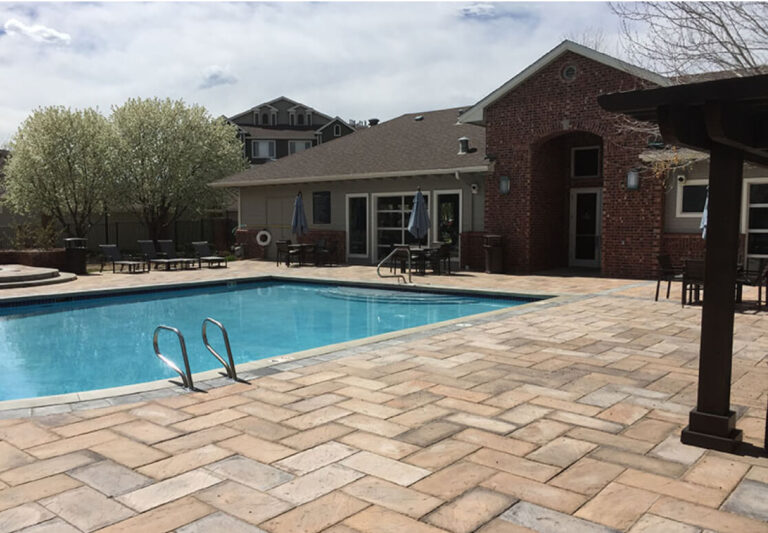 Outdoor swimming pool with metal ladder, surrounded by a paved patio, lounge chairs, and a brick building in the background under a partly cloudy sky.