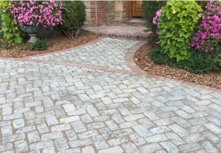 A stone paver walkway with a brick border leads to a front door, surrounded by green shrubs and blooming pink flowers.