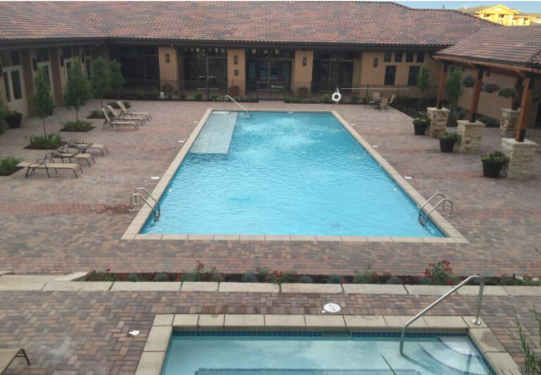 Rectangular outdoor swimming pool surrounded by lounge chairs and potted plants on a brick patio, with a building in the background and a hot tub in the foreground.