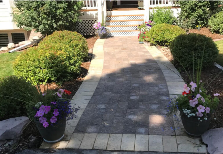 A paved walkway with two flower pots on either side leads to the steps of a house, surrounded by trimmed bushes and garden beds.
