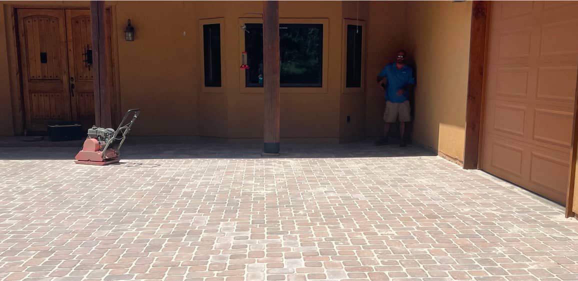 A man stands in the shade near a garage and wooden door, with a paving machine on a newly installed brick driveway.