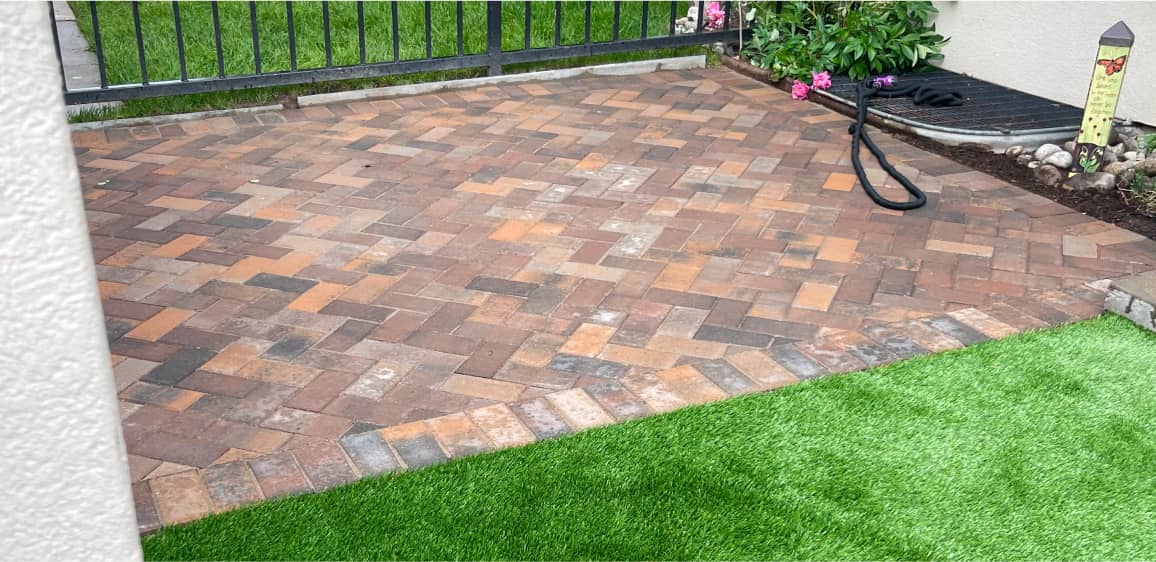 A patio area with interlocking brown and orange pavers in a herringbone pattern is bordered by artificial green grass, a garden bed, and a metal fence.